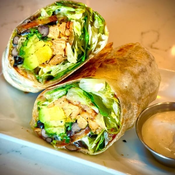 Two halves of a vegan burrito filled with lettuce, avocado, black beans, and plant-based protein, with a side of sauce.