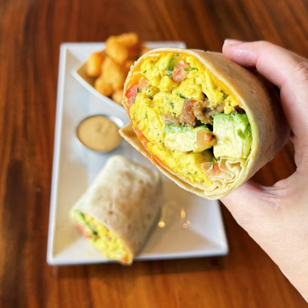 A hand holds a vegan breakfast burrito filled with scrambled eggs, avocado, and tomatoes, with a side of tater tots and sauce.