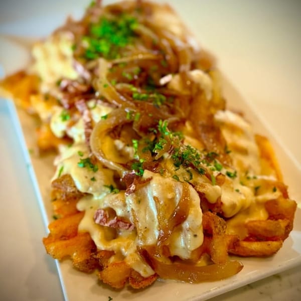 Vegan waffle fries topped with melted cheese, caramelized onions, and fresh parsley on a white plate.