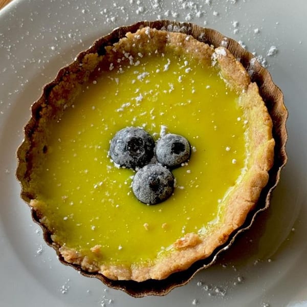 A vegan lemon tart with three blueberries and powdered sugar on a white plate.
