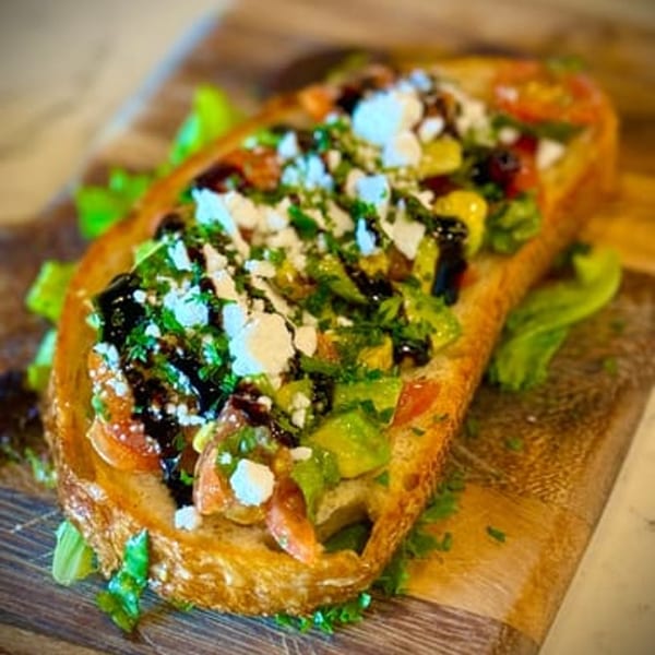 Ocado Restaurant's vegan avocado toast with tomatoes, feta, herbs, and balsamic glaze on a wooden board.