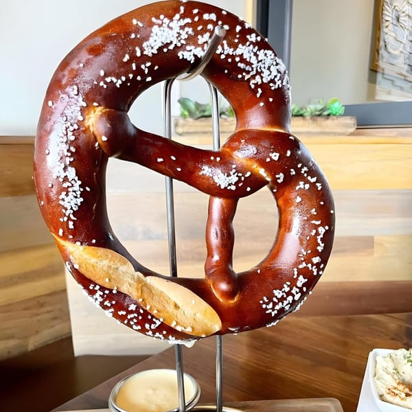 A large, salted vegan pretzel hangs from a metal stand with dipping sauces below.