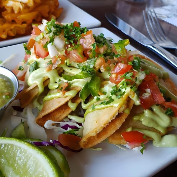 Crispy vegan taquitos topped with fresh salsa, green sauce, and a side of lime wedges.
