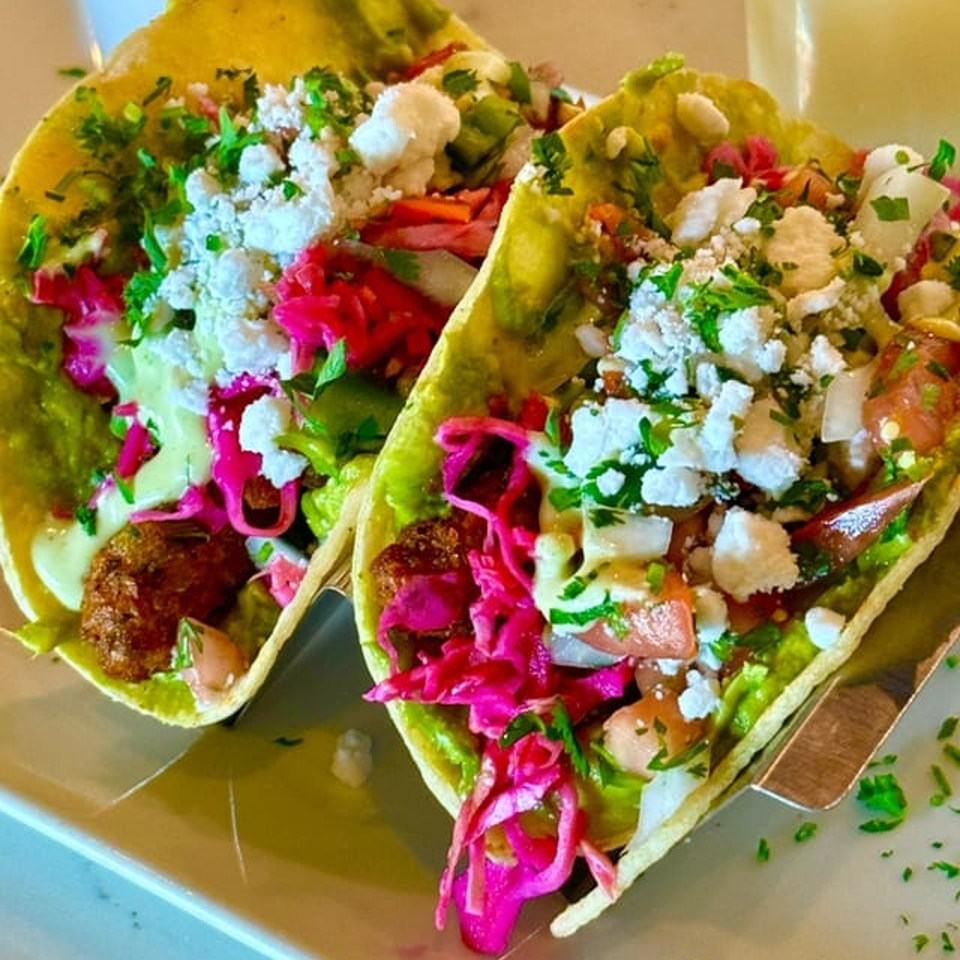 Two vibrant vegan tacos with falafel, guacamole, pickled cabbage, and crumbled cheese on a white plate.
